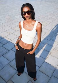 Valkoinen ribbattu crop-top, jossa on napit, yhdistetty löysiin mustiin leveisiin housuihin. Mallilla on päällään ylisuuria tummia aurinkolaseja ja hopeisia rannekoruja.