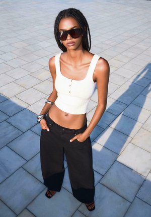 Weißes, geripptes Crop-Top mit Knöpfen, kombiniert mit lockeren, schwarzen, weiten Hosen. Das Modell trägt übergroße, dunkle Sonnenbrillen und silberne Armreifen.