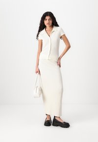 Top tricotat alb cu textură, cu mâneci scurte, asociat cu o fustă lungă până la podea, de aceeași nuanță. Modelul poartă o geantă albă mică și pantofi cu platformă negri.