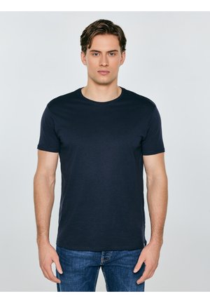 Młody mężczyzna o krótkich brązowych włosach, ubrany w prostą czarną koszulkę z krótkim rękawem i niebieskie dżinsy, stojący na tle jasnoszarego tła.