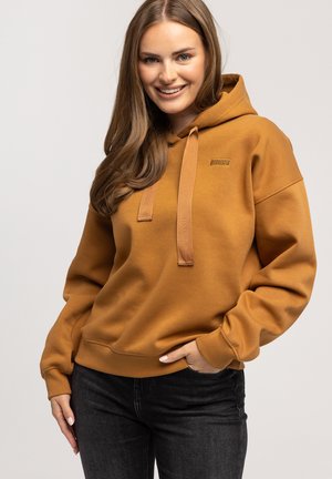 Mustardowy sweatshirt z kapturem, z przednią kieszenią i naszywką z logo, w komplecie z ciemnymi dżinsami; miękki materiał i ściągacze na rękawach.