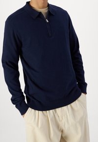 Pull-over bleu marine à manches longues avec col zippé, texture tricotée, et poignets et ourlet côtelés, associé à un pantalon beige clair.