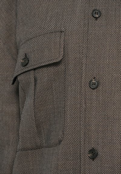 Primer plano de un bolsillo de una chaqueta marrón texturizada con una solapa abotonada y tres botones negros visibles en la parte frontal.