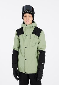Grüne isolierte Jacke mit schwarzen Akzenten, mit Reißverschluss versehene Brusttasche, verstellbare Bündchen und eine schützende Kapuze; getragen mit Handschuhen und Schutzbrille.