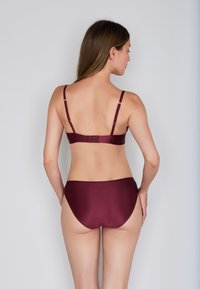 Frau mit langen braunen Haaren, die einen dunkelroten, gerippten Bikini trägt, von hinten vor einem schlichten hellgrauen Hintergrund gezeigt.