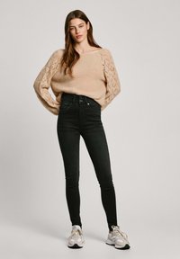 Suéter de punto beige con detalles de encaje, combinado con jeans negros de cintura alta y zapatillas deportivas beige. Tonos neutros sencillos en toda la combinación.