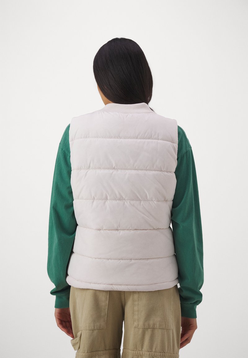 Gepolsterte Weste in hellbeige mit horizontalem Steppmuster, ausgestattet mit einem gerippten Kragen und getragen über einem grünen Langarmshirt.