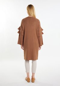 Lange, loszittende bruine cardigan met gelaagde ruches aan de mouwen, gecombineerd met lichtbeige broek en lichtbruine instap schoenen, gezien vanuit de achterkant.