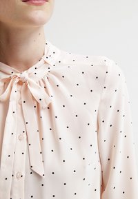 Blouse rose clair à petits pois noirs, dotée d'un nœud papillon noué au col, fermeture par boutons, et tissu doux et lisse.