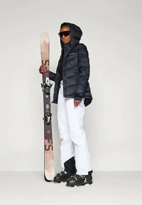 Jachetă puffer neagră, pantaloni de schi albi, cizme de schi închise la culoare, ținând schiuri cu un design maro și roz. Detalii despre ochelari de soare și jacheta cu glugă sunt vizibile.