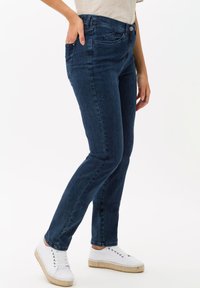 Jean en denim bleu foncé à coupe slim, avec cinq poches et un bouton de fermeture. Portés avec des espadrilles blanches. Texture lisse, design classique.