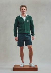 Groene gestructureerde cardigan met logo, witte blouse met kraag, donkere shorts en witte perforerende schoenen, staand op een houten voetstuk.