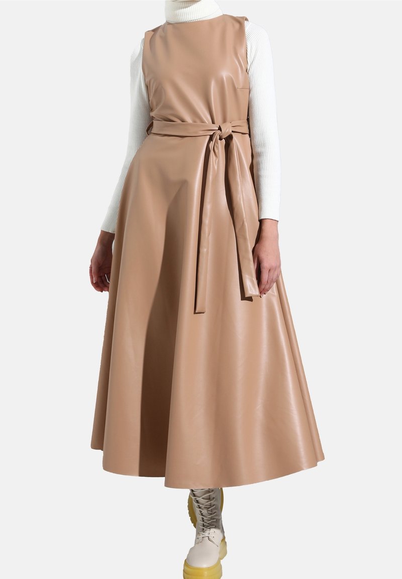 Robe midi sans manches en cuir synthétique couleur beige avec ceinture nouée à la taille, portée sur un pull à col roulé blanc côtelé et des bottines beige à lacets.