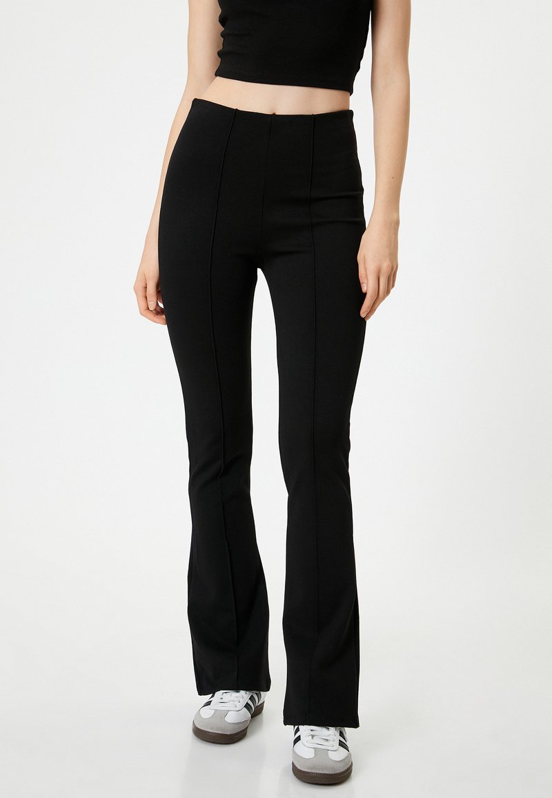 Pantalons évasés noirs en tissu extensible. Possède une taille haute avec des coutures visibles et une texture lisse. Portés avec des baskets blanches.