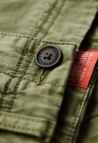 Tissu en coton vert avec une texture lisse, présentant un bouton marron foncé étiqueté "SUPERDRY" et une étiquette de marque rouge visible.