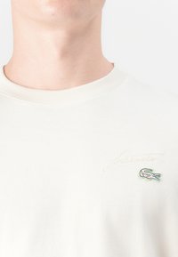 T-shirt en coton crème, avec un col rond, arborant un script "Lacoste" brodé et un petit logo crocodile vert sur la poitrine.