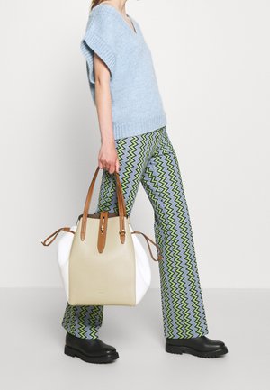 Personne portant un pull bleu clair à manches courtes, un pantalon à motif zigzag, des bottes noires, tenant un grand sac à main beige et blanc avec des anses marron.