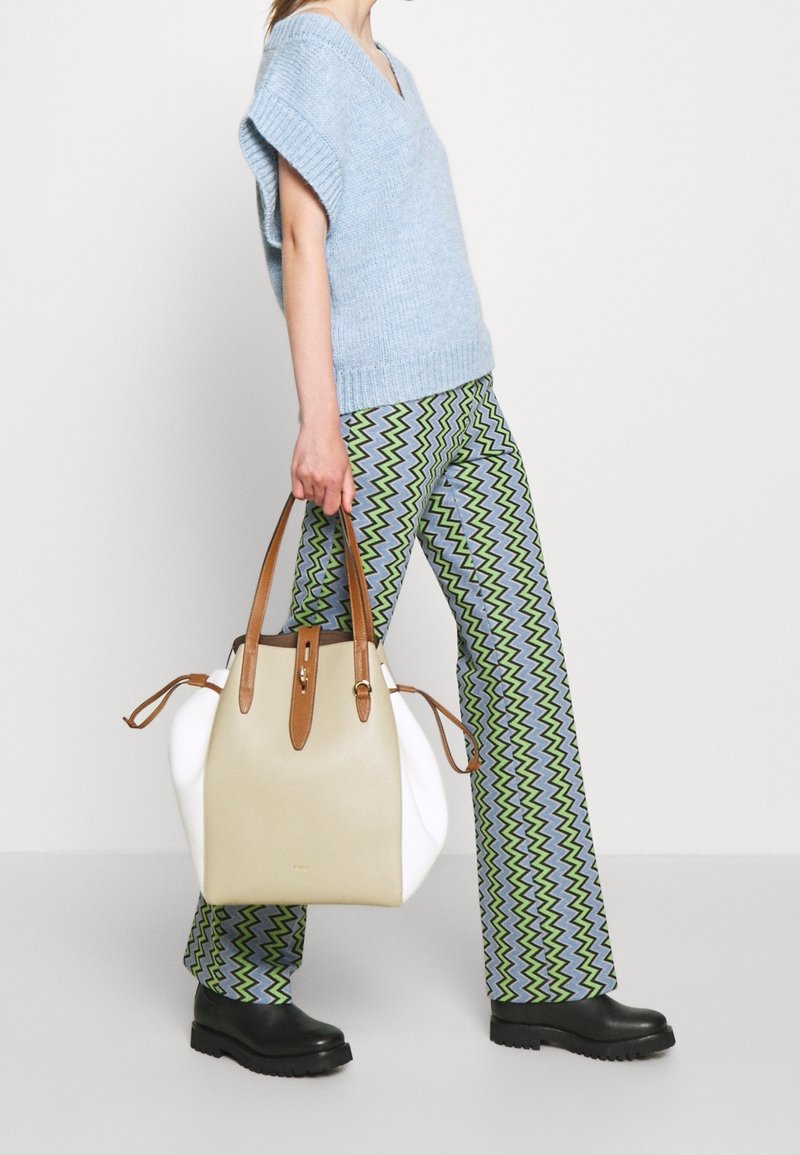 Personne portant un pull bleu clair à manches courtes, un pantalon à motif zigzag, des bottes noires, tenant un grand sac à main beige et blanc avec des anses marron.