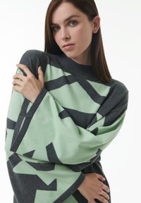Grauer und mintgrüner Oversize-Pullover aus weichem Stoff, mit einem markanten geometrischen Muster und weiten Ärmeln sowie einem ribbed Halsauschnitt.