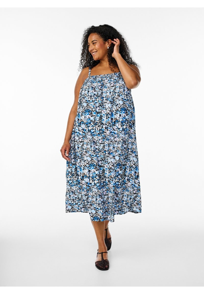 Femme aux cheveux bouclés portant une robe midi sans manches bleue à fleurs et des chaussures noires à bride à la cheville, souriante et se touchant les cheveux.