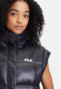 Svart pufferväst med hög krage, korta ärmar och glänsande yta. Har en broderad vit "FILA"-logotyp på framsidan.