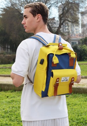 Rucksack aus gelbem und blauem Stoff, mit Lederakzenten, verstellbaren Trägern, Fronttasche und Druckknopfverschlüssen. Hat ein robustes Design.