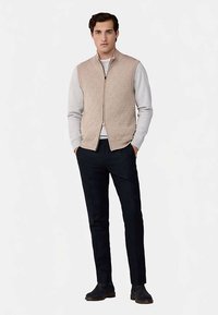 Beige gesteppte Weste mit einem Reißverschluss vorne, getragen über einem grauen Hemd. Kombiniert mit dunkelblauen Hosen und schwarzen Schuhen, besticht das Outfit durch ein schlichtes Design.