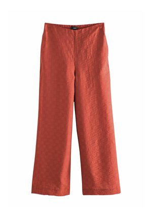 Jeune femme aux longs cheveux blonds portant un haut sans manches à col halter rouge rouille et un pantalon large assorti, debout contre un mur beige.