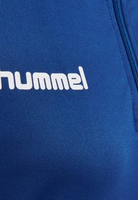 Modrá sportovní bunda s hladkou texturou, plným zipem a výrazným bílým logem "hummel" na přední straně.