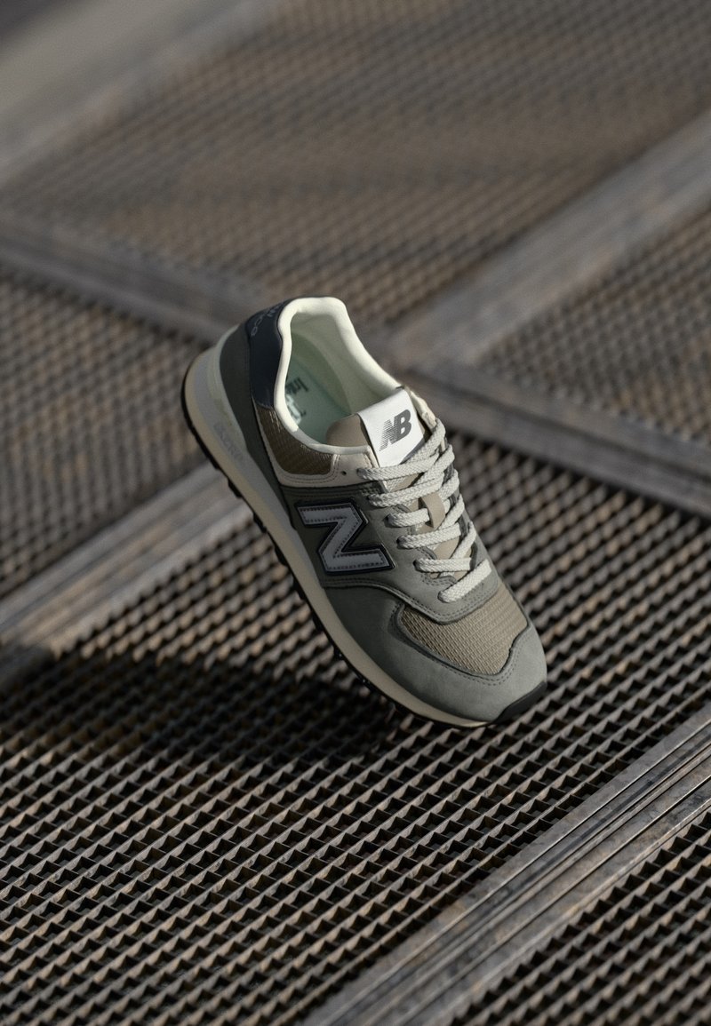 Baskets New Balance grises et blanches avec lacets sur une surface de grille métallique texturée, éclairées par la lumière naturelle et projetant une ombre.