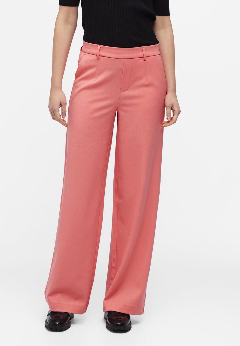 Pantalones de pierna ancha en tejido de color coral rosa con una textura suave, que presentan una cinturilla plana y pliegues en el frente. Llevados con zapatos negros.