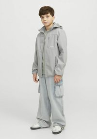 Grijze lichte jas met een capuchon, voorzakken met rits en merklogo. Gecombineerd met losse blauwe cargobroeken en sneakers met witte accenten.