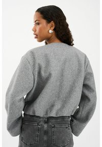 Kobieta z kręconymi włosami, nosząca duże srebrne kolczyki, szary top z długim rękawem i czarne dżinsy z wysokim stanem, stojąca bokiem na jednolitym tle.