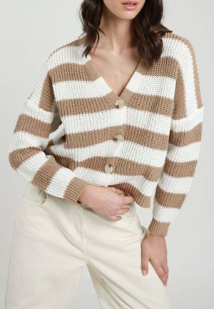 Donna che indossa un cardigan lavorato a maglia a righe beige e bianche con bottoni e pantaloni bianco sporco, posando con una mano sulla vita e l'altra sulla coscia.