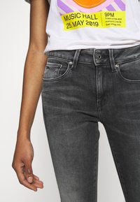 Vyprané černé džínové kalhoty s přiléhavým střihem, pěti kapsami a kovovým zapínáním na knoflík. Tkanina má mírný lesk a hladkou texturu.