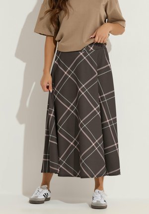 Kobieta ubrana w brązową, kratowaną, midi spódnicę w kształcie litery A, beżową bluzkę z krótkim rękawem oraz białe trampki z czarnymi paskami, stojąca przy gładkiej ścianie.