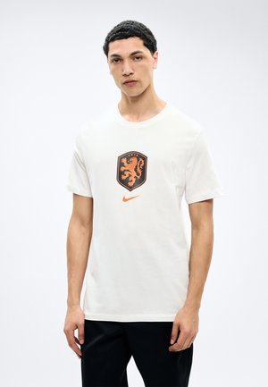 Mann iført hvit T-skjorte med svart og oransje løveemblem og Nike-logo, stående mot enkel lys bakgrunn.