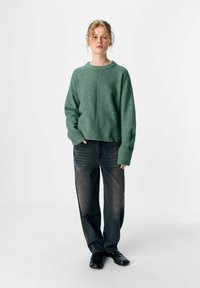 Jongere met krullend haar, gekleed in een groene trui, losse zwarte jeans en zwarte schoenen, staand tegen een eenvoudige lichte achtergrond.