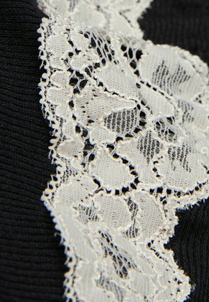 Svart tyg med en bred, intrikat vit spetsfåll som har blommönster längs ena sidan och en texturerad stickad bakgrund.