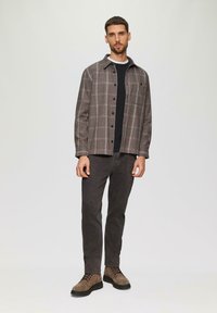 Geruite button-up shirt in bruin en grijs, gedragen over een zwarte top. Slim-fit donkere broek en bruine laarzen maken de outfit compleet.