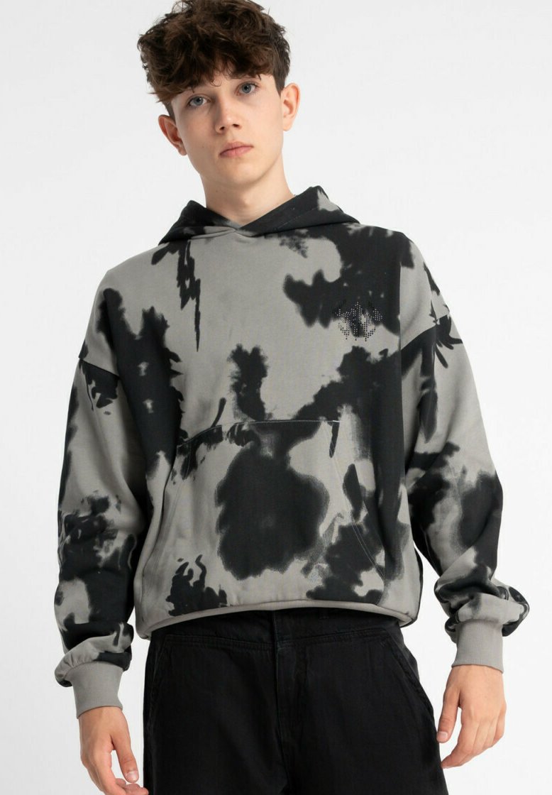 Młody mężczyzna o kręconych włosach, ubrany w szaro-czarny hoodie w stylu tie-dye i czarne spodnie, stojący na tle białej ściany.