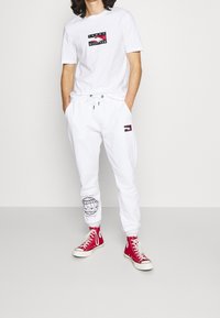 Vit bomullströja med en svart och röd logotyp, tillsammans med vita sweatpants med en grafik på vänster ben och röda högskor.