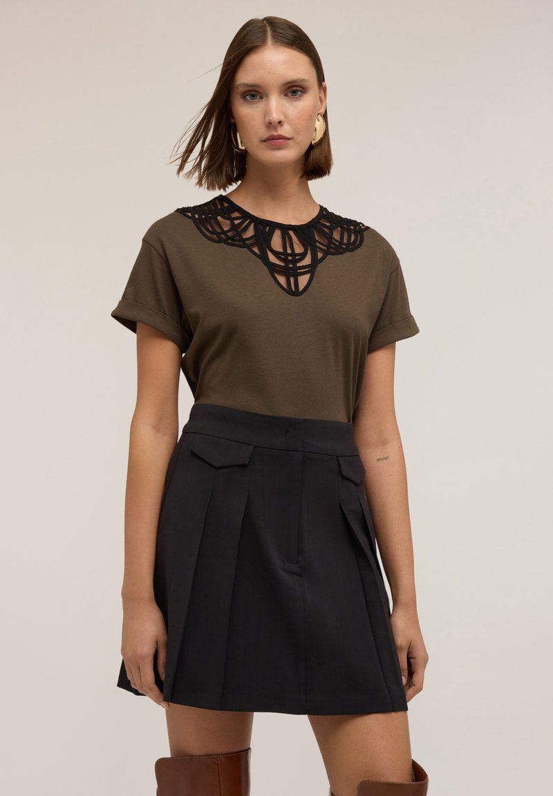 Olivově zelené tričko s černými vyřezávanými krajkovými detaily kolem výstřihu, kombinované s černou skládací sukní s postranními kapsami.