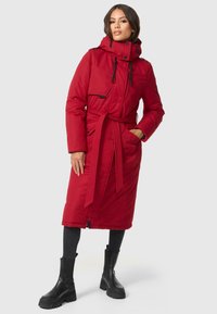 Manteau long rembourré rouge avec une taille cintrée, poches zippées, col montant et capuche avec cordon de serrage, associé à des bottines noires et un jean foncé.