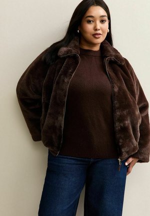 Frau in einer dunkelbraunen Kunstpelzjacke über einem passenden Pullover und blauen Jeans, die vor einem einfarbigen beigen Hintergrund steht.