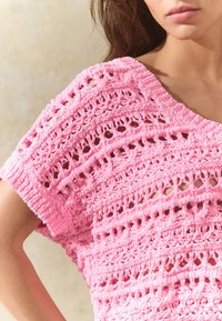 Pink crochet top featuring an open knit design with a loose, wide neckline and short sleeves, displaying intricate patterns throughout.
