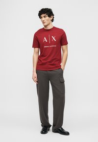 Νεαρός άνδρας με σγουρά μαλλιά, φορώντας κόκκινο t-shirt Armani Exchange και σκούρο γκρι παντελόνι cargo, στέκεται με το ένα χέρι στην τσέπη του.