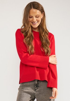 Femme souriante aux longs cheveux ondulés, portant un pull rouge à manches longues et un jean gris, debout avec un bras croisé sur l'autre.