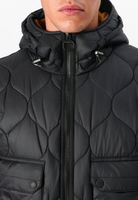 Casaco puffer preto acolchoado com capuz, fecho frontal e dois bolsos no peito com fecho de pressão. Textura suave e design minimalista.