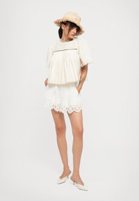 Woman wearing a white lace blouse and shorts, straw hat, and white kitten heel mules, standing with hands in pockets against a plain background.